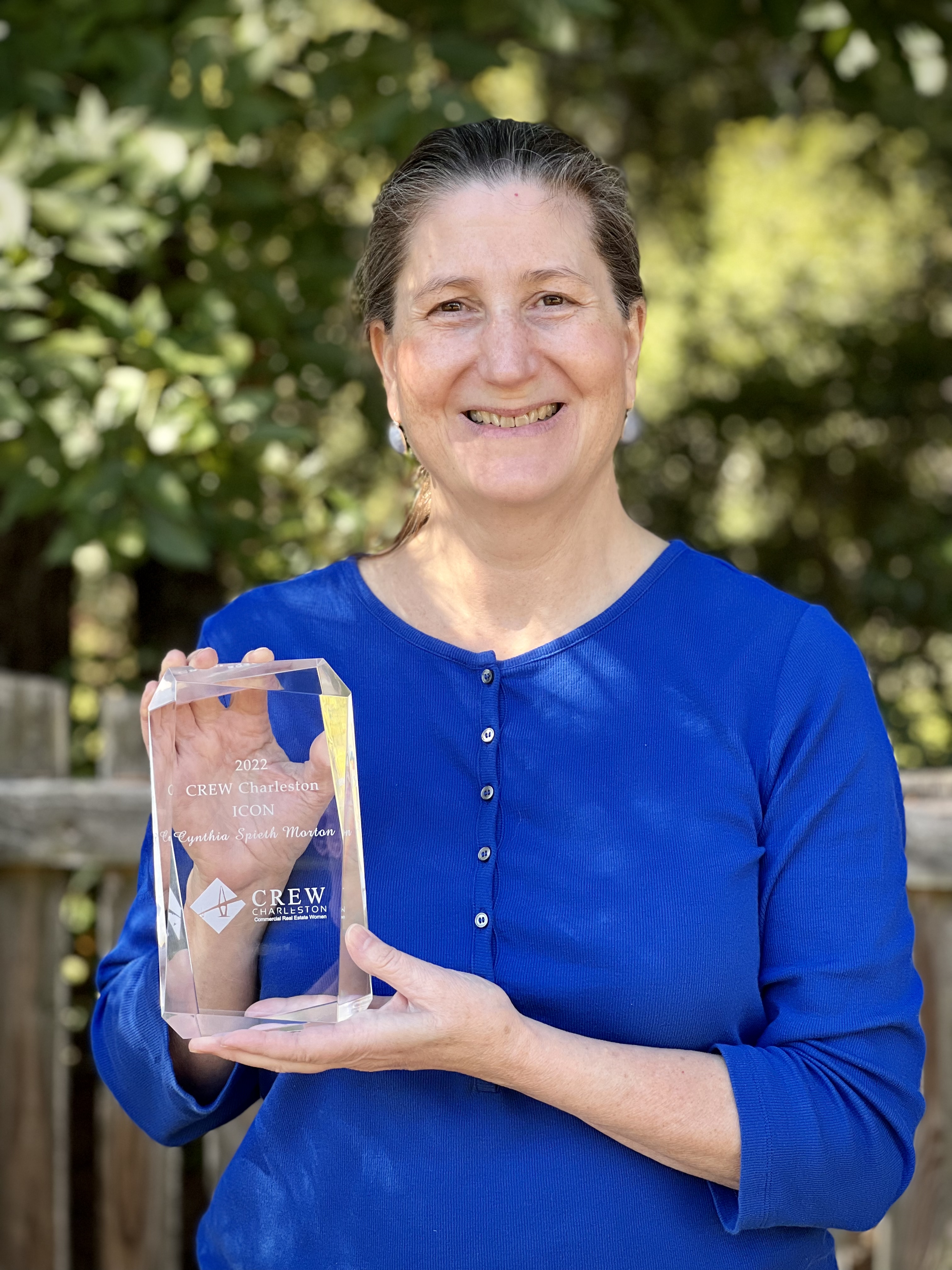 Cynthia Spieth Morton holds a plaque for her 2022 CREW (Commercial Real Estate Women) Charleston Icon Award. She is wearing a blue shirt and stands in front of a green, natural background.