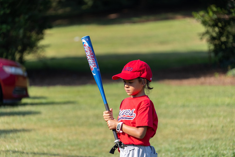 A child with a shirt and hat that read "Braves" looks at a tee ball bat.