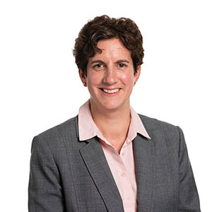 Professional woman smiling in a business portrait with a grey suit jacket and light pink shirt, against a neutral background.