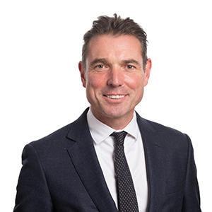 Professional businessman wearing a dark suit with a white shirt and patterned tie, smiling confidently against a white background.