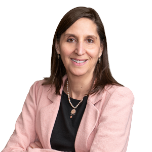 Smiling professional woman wearing a pink blazer over a black top, with a pendant necklace, against a white background.