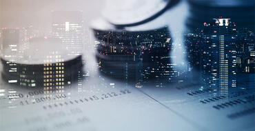Three piles of coins lay on top of financial records, while offices are illuminated in tall corporate buildings in the background. From Shutterstock, ID 522559909
