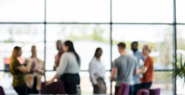 Blurred image of a diverse group of people gathered inside an office space with large windows during a casual meeting or networking event.