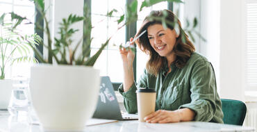 Smiling woman working from home at her minimalistic desk space, with a laptop, coffee cup, and houseplants in a bright, airy room.