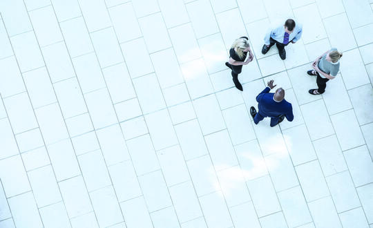 Aerial view of four business professionals in a casual meeting on a white tiled floor, highlighting teamwork, corporate environment, and strategic discussion.