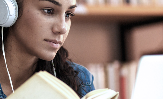 Focused young woman studying with headphones reading a book in front of her laptop in a home environment