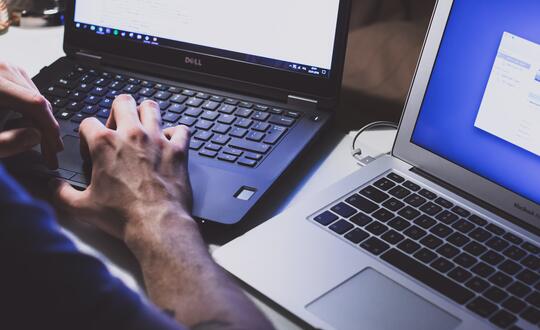 Person typing on a Dell laptop while multitasking with a second MacBook, indicating productivity and efficient workflow in an office environment.