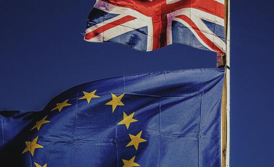 British Union Jack flag fluttering above a European Union flag against a clear blue sky.