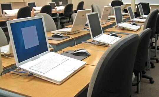 Laptop with a blue screen error on a desk in a computer lab with multiple workstations and desktop PCs.