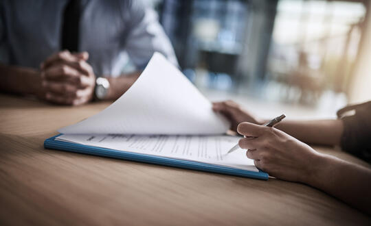 Two people are sitting at a table. A man is clasping his hands and a woman is holding a pen, looking over a file folder with paperwork laying on top.
