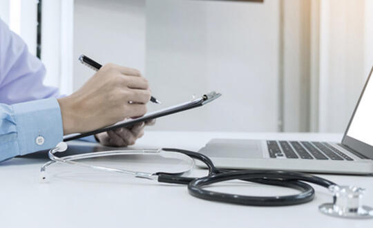 Healthcare professional writing notes with a stethoscope and laptop on a desk, depicting medical consultation or patient record management in a clinical practice setting.