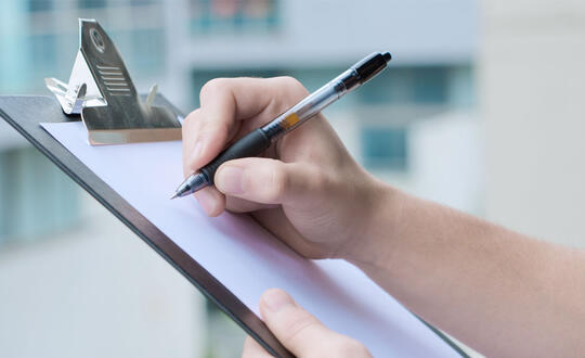 Individual holding a pen up to write on a piece of paper on a clip board - Shutterstock 110625482