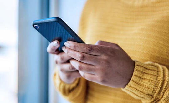 A woman in a yellow sweater looks down at a cell phone she holds in her hands.