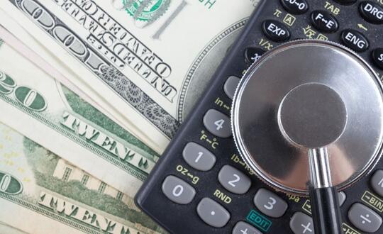 A stethoscope sits on top of a calculator in front of a background of U.S. money