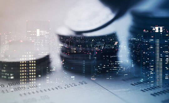 Three piles of coins lay on top of financial records, while offices are illuminated in tall corporate buildings in the background. From Shutterstock, ID 522559909