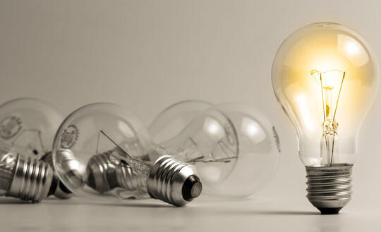 A lit lightbulb stands beside a pile of darkened lightbulbs