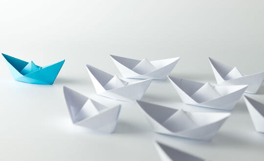 A group of white origami boats with a blue origami boat leading at the front - Shutterstock 318124145