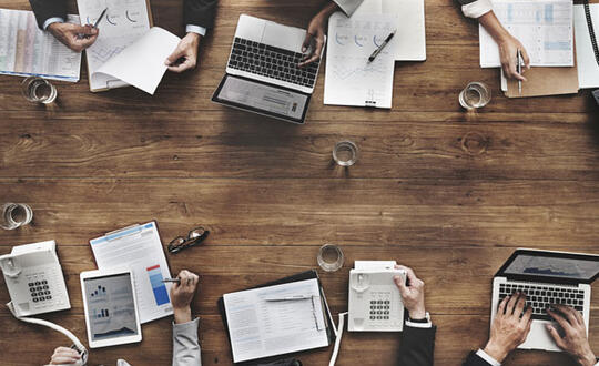 Six businesspeople look at various reports and laptops during a business meeting - Shutterstock 374801434