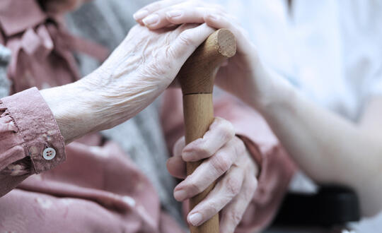 A caregiver holds the hand of an elderly man as he grips his cane.