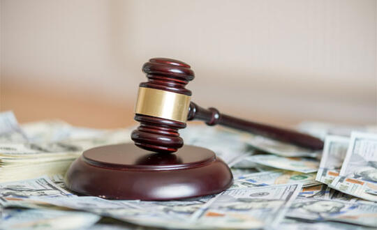A gavel lays on top of a pile of one hundred dollar bills in U.S. currency.