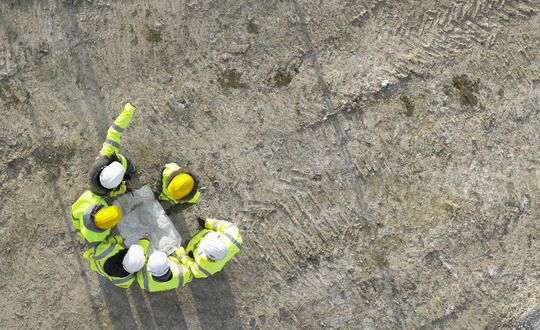Group of construction workers in high-visibility jackets having a meeting on a building site with safety helmets visible, top view on a textured muddy ground background.
