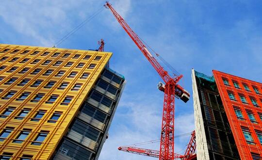 Construction crane between two modern buildings with vibrant yellow and red façades against a clear blue sky, depicting urban development and architecture in a bustling city centre.