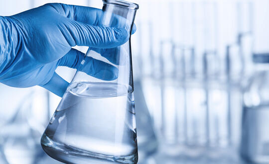 A gloved hand holds a beaker filled with clear liquid, with more beakers lined up behind.
