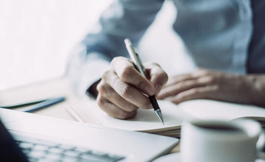 Close-up of a person's hands writing in a notebook with a pen, with a blurred coffee cup in the foreground, suggesting a focus on business planning or creative writing in a relaxed environment.