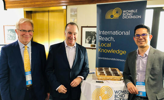 Attorneys Barlow Keener, Marty Stern, and Bob Silverman stand in front of a sign that reads, "Womble Bond Dickinson: International Reach, Local Knowledge."