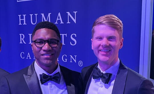 Attorneys Nick Hawkins and Mason Freeman stand in front of a blue banner that reads in white text, "Human Rights Campaign".
