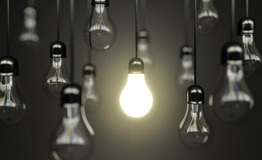 Several lightbulbs hang in front of a black backdrop. Only one lightbulb is illuminated. Source: Shutterstock