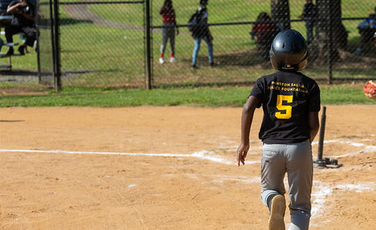 A child runs to home base during a tee ball game. The back of his shirt reads, "Winston-Salem Police Foundation."