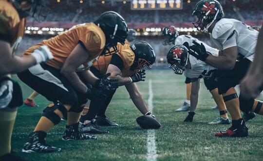 American football players crouched in a line of scrimmage pre-match on a floodlit field, showcasing teamwork, sports gear, and competitive atmosphere.
