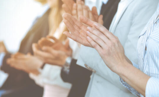 Group of professionals clapping hands in a bright office environment, signifying teamwork and success at a corporate event or business meeting