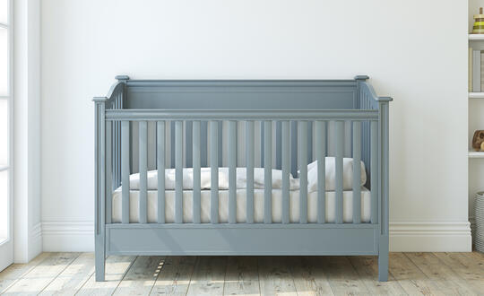 A blue and gray crib sits inside a room, beside baby toys and an open window with light coming in.