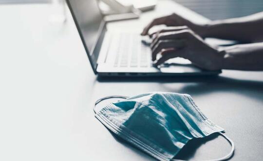 Person typing on a laptop with a disposable face mask on the desk, implying remote work during a pandemic.