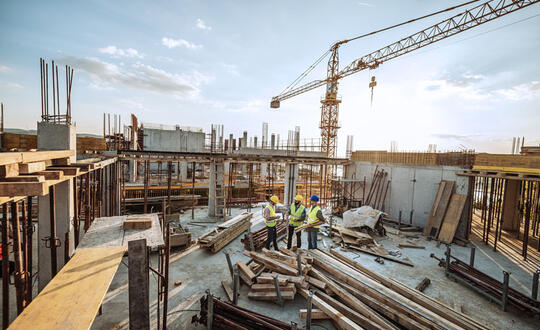 Construction workers discussing plans on a building site with scaffolding, steel reinforcements, and a crane in the background during late afternoon.