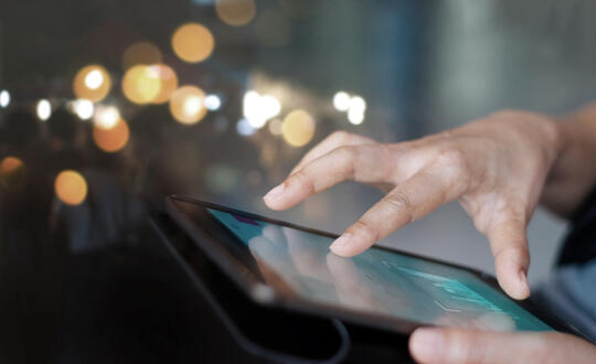 Person using a digital tablet with touchscreen interface in an urban setting with bokeh lights in the background.