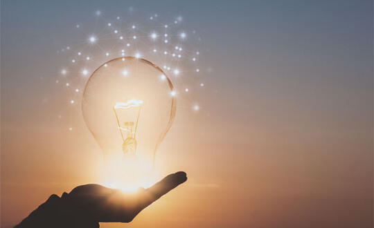 A person cups their hand under a lighted lightbulb, in front of a sunset. 