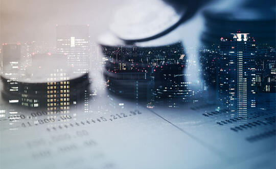 Three piles of coins lay on top of financial records, while offices are illuminated in tall corporate buildings in the background. From Shutterstock, ID 522559909