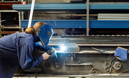 Female welder utilising welding equipment to fabricate metalwork in an industrial workshop-setting, wearing safety gear and workwear.