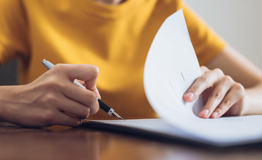 Person in a yellow shirt writing notes on a white paper at a wooden desk.