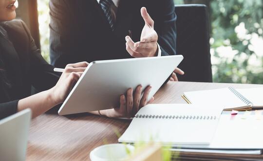 Professional business meeting with digital tablet, spiral notebook, and pen on wooden table, team discussion in modern office.