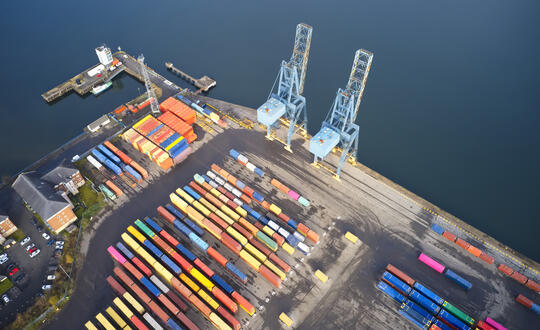 Aerial view of a bustling cargo port with colourful shipping containers arranged in neat rows, large cranes ready to load and unload goods, and a cargo ship docked at the quayside, showcasing the vibrant activity of maritime logistics and international trade.