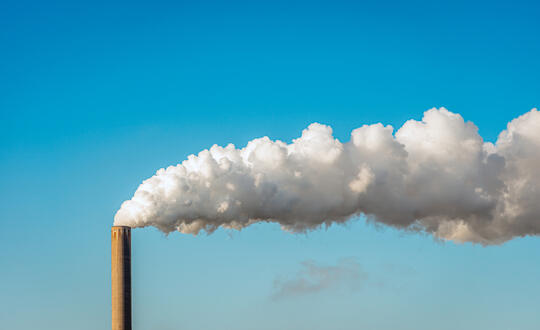 Industrial chimney emitting white smoke against a blue sky, symbolising air pollution and environmental issues in manufacturing sectors.