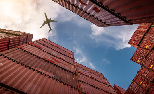 View from below of stacked shipping containers with a plane flying overhead against a blue sky with clouds, symbolising global trade and transport logistics.