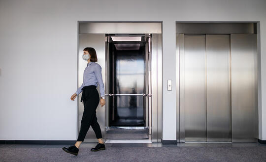 Woman wearing a mask walking past an open lift in a modern office building.