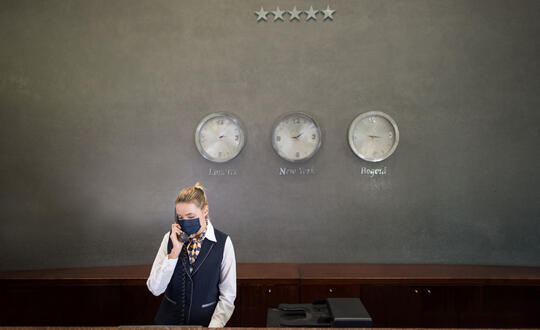 Businesswoman wearing a mask standing before a wall with clocks showing different time zones, including London, New York, and Beijing, symbolising global connectivity and international business operations.