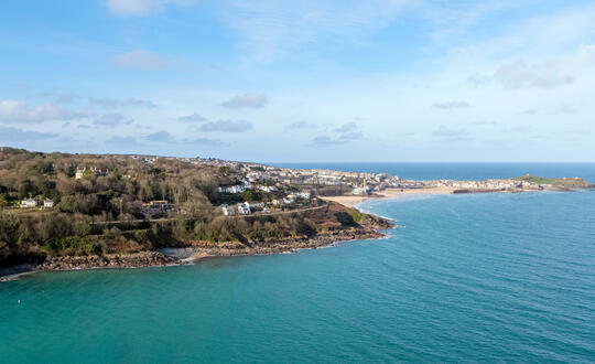 Aerial view of a picturesque British seaside town with lush greenery, coastal cliffs, and azure waters under a clear blue sky.