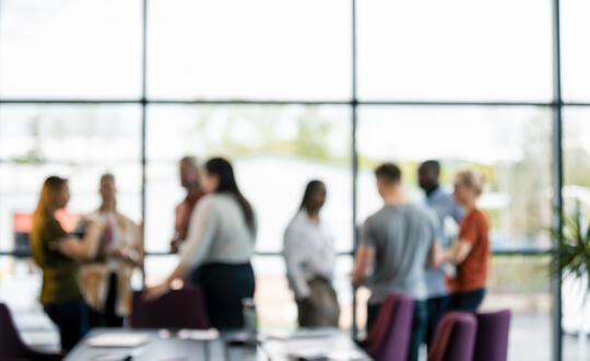 Blurred image of a diverse group of people gathered inside an office space with large windows during a casual meeting or networking event.